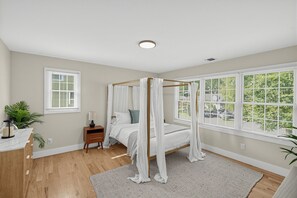 A bedroom with a four-poster bed, a bedside table with a lamp, a large window with blinds, and a view of greenery outside. - Enjoy Quiet Nights With A Fireplace In Bellport 3 Bedroom Home (Bellport)