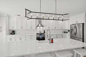 A modern kitchen with white cabinetry, stainless steel appliances, a central island with bar stools, and pendant lighting. - Enjoy Quiet Nights With A Fireplace In Bellport 3 Bedroom Home (Bellport)