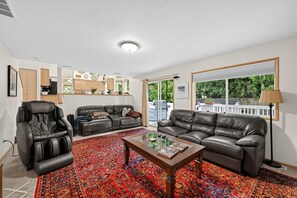 Ein Wohnzimmer mit einer Couch, einem Liegesessel, einem Couchtisch und einem großen Fenster mit Blick auf Grünflächen.