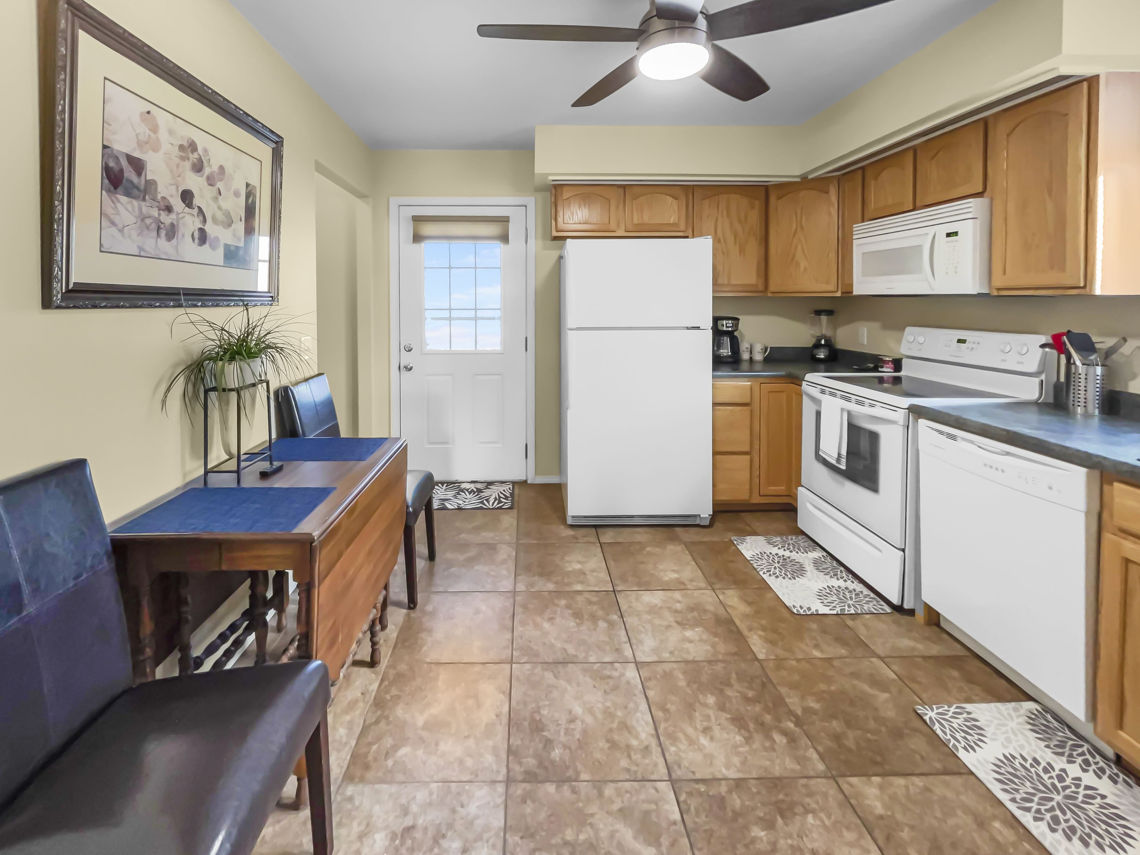 A compact kitchen with a refrigerator, microwave, stove, and oven. There is a dining table with chairs and a ceiling fan.