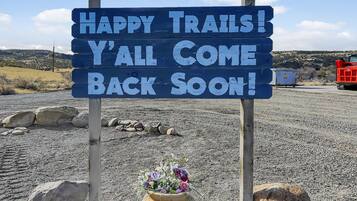 A sign with the text 'HAPPY TRAILS! Y'ALL COME BACK SOON!' in a rural setting with a gravel ground and a clear sky.