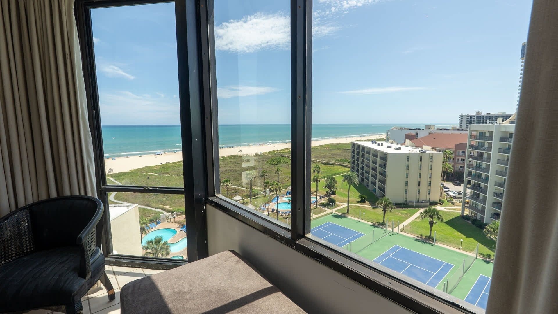 Ein Zimmer mit großem Fenster mit Blick auf einen Strand, einen Pool und einen Tennisplatz.