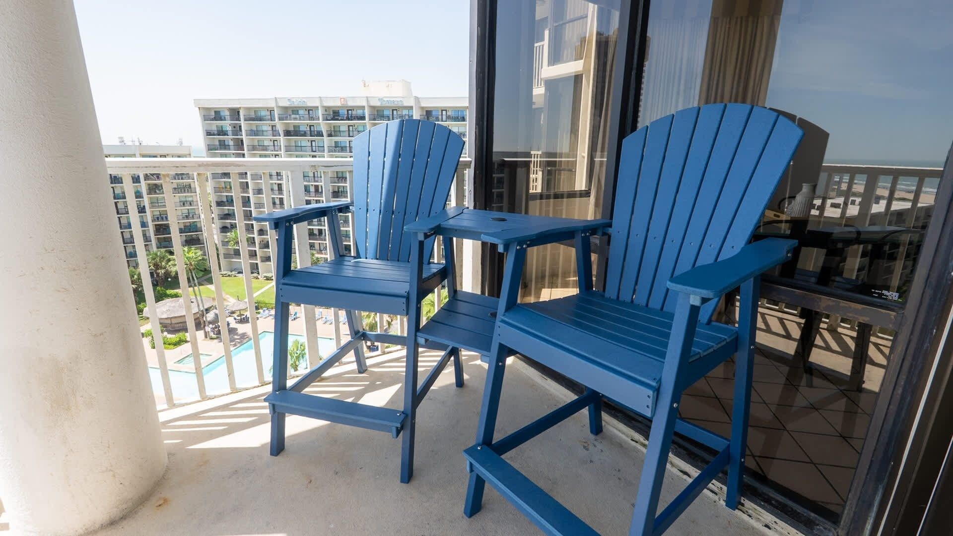 A balcony with two blue chairs and a small table, overlooking a pool and buildings.