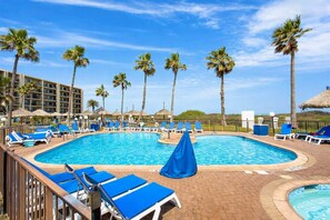 Piscina con sillas reclinables azules y palmeras, rodeada por un edificio de hotel.