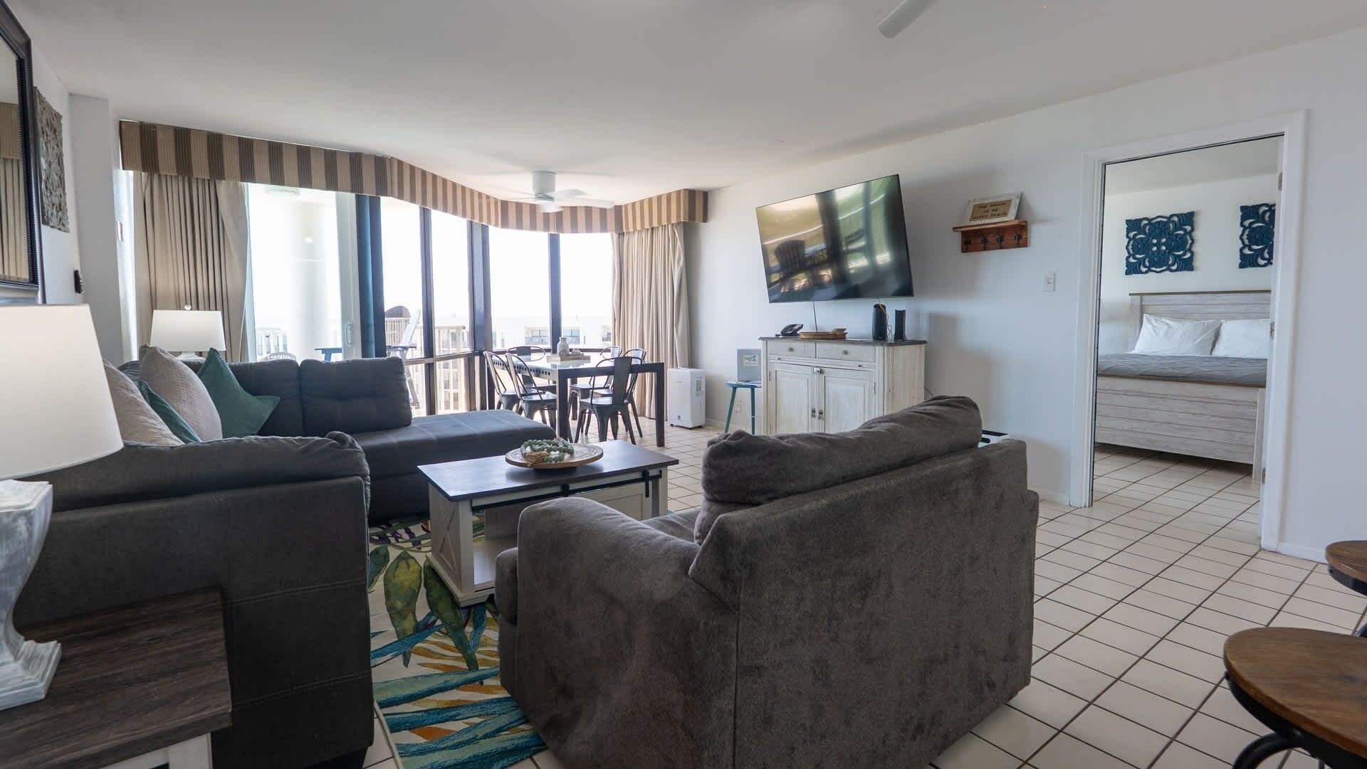 A modern living room with a sofa, armchairs, a dining table, and a TV mounted on the wall.