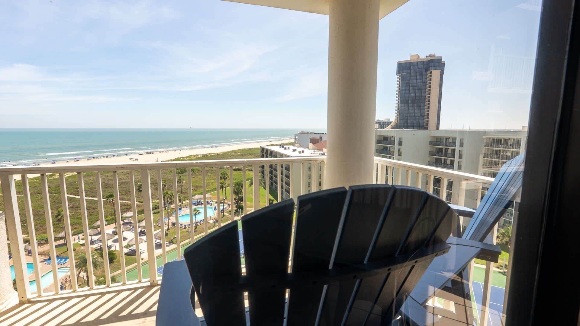 A balcony with a view of a beach and ocean, a pool area, and high-rise buildings in the background.