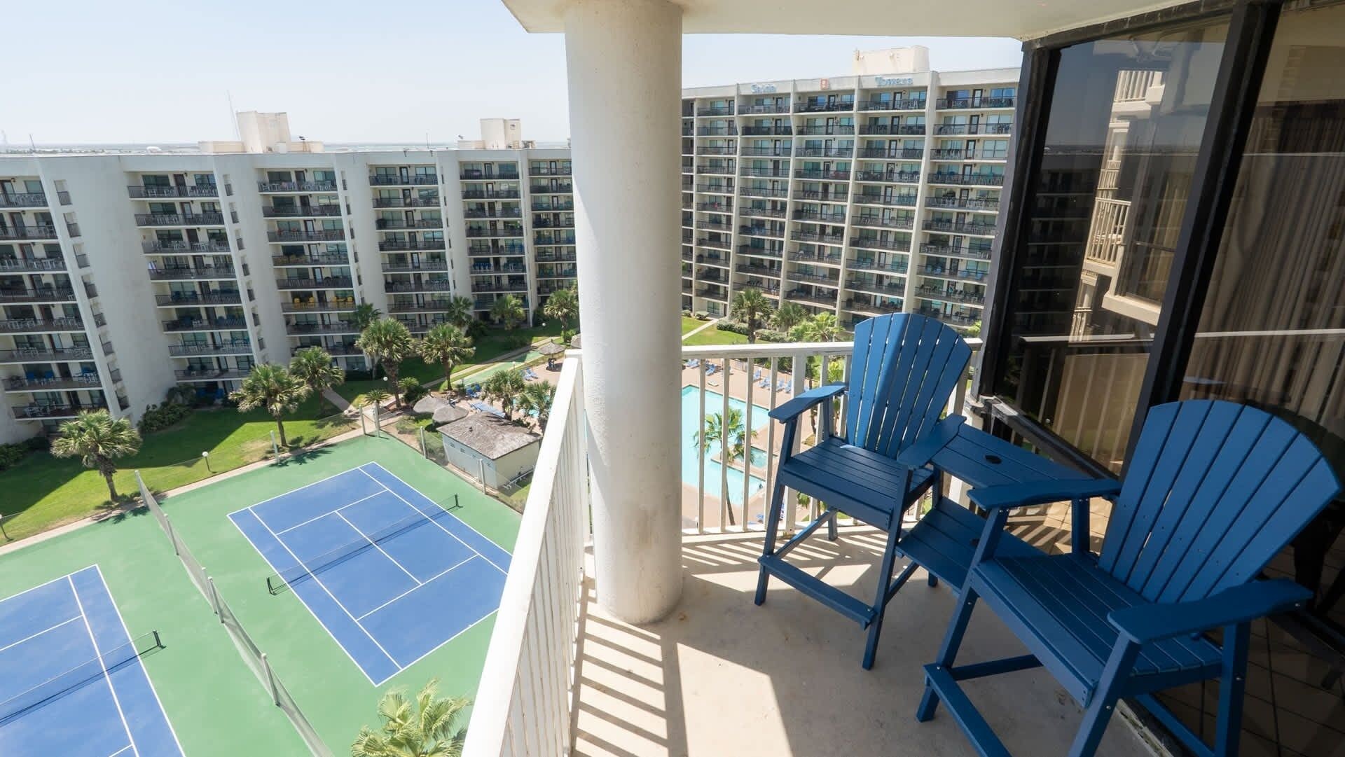 A balcony with blue Adirondack chairs, a tennis court, and a view of a hotel building with multiple balconies.