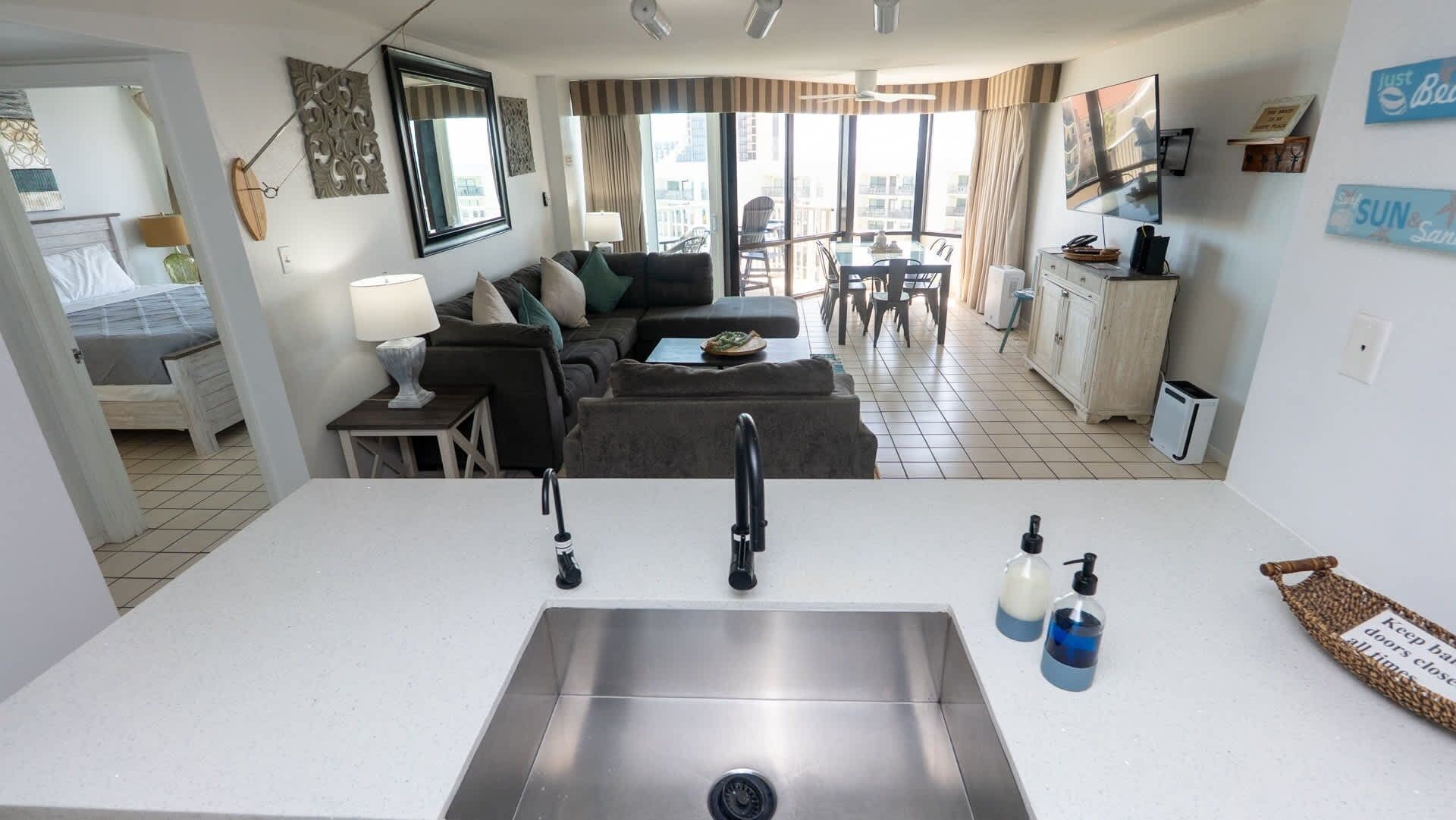 A modern kitchen with a stainless steel sink, a dining area with a table and chairs, a living room with a sofa and a TV, and a balcony with a view of the city.