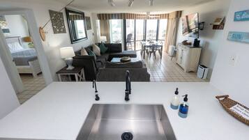 A modern kitchen with a stainless steel sink, a dining area with a table and chairs, a living room with a sofa and a TV, and a balcony with a view of the city.