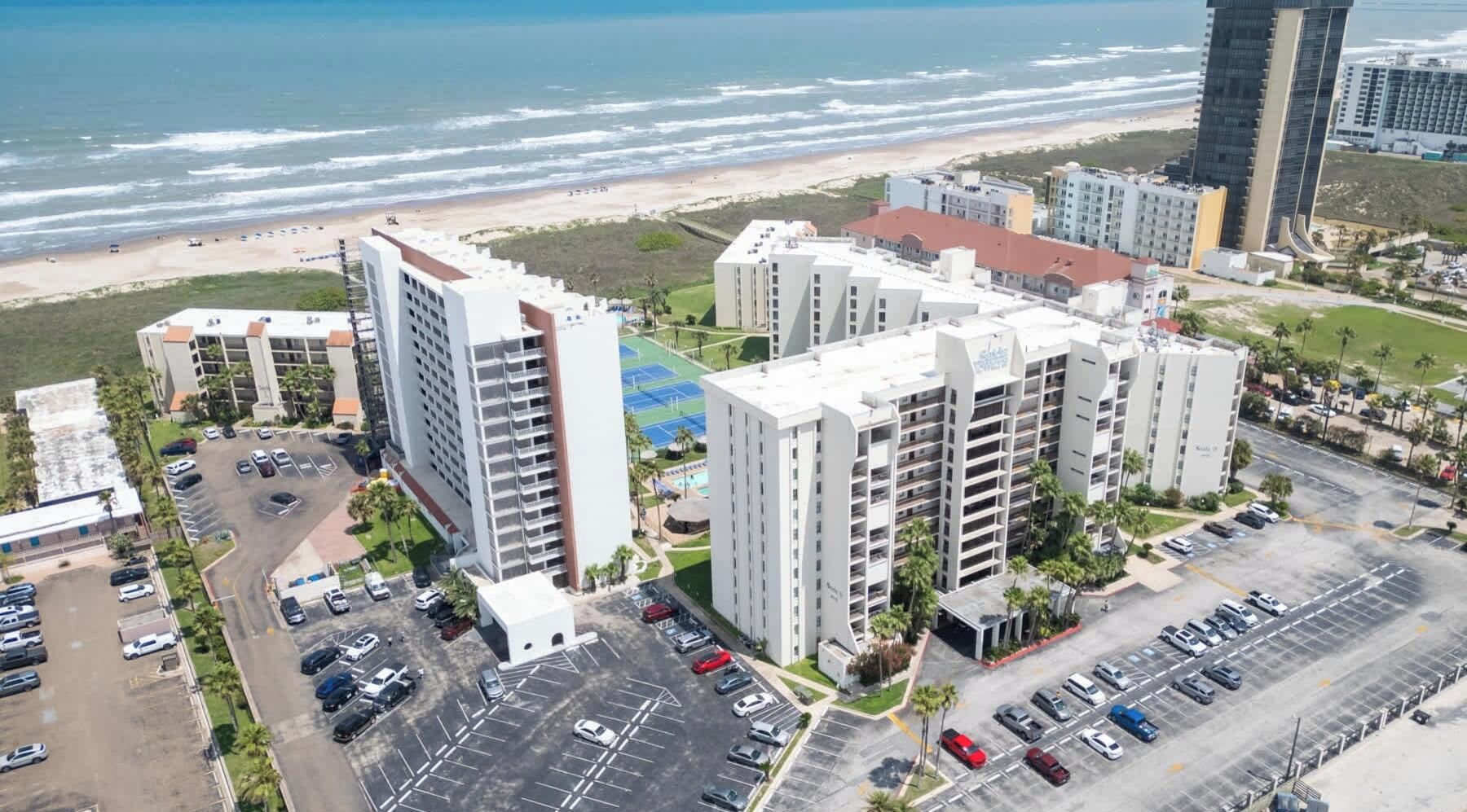 A high-rise hotel with multiple floors, a parking lot, and a beach view.