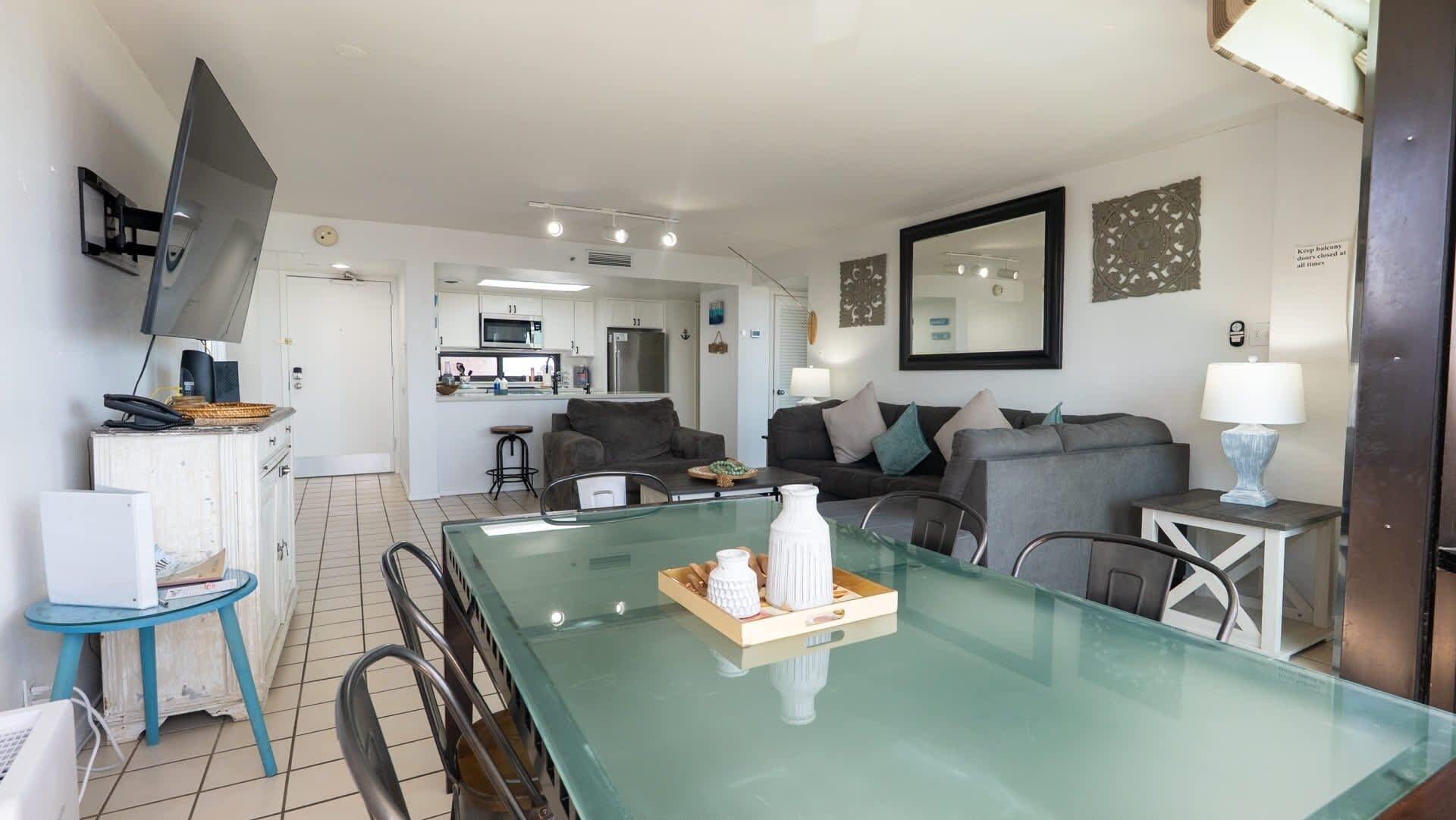 A modern living room with a glass dining table, grey sofas, and a kitchen in the background.