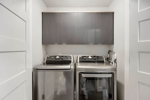 A compact laundry room with two washing machines and a dryer, featuring a modern design and a grey cabinet above. - Shangri-la 102 3 Bedroom Apts (Heber City)