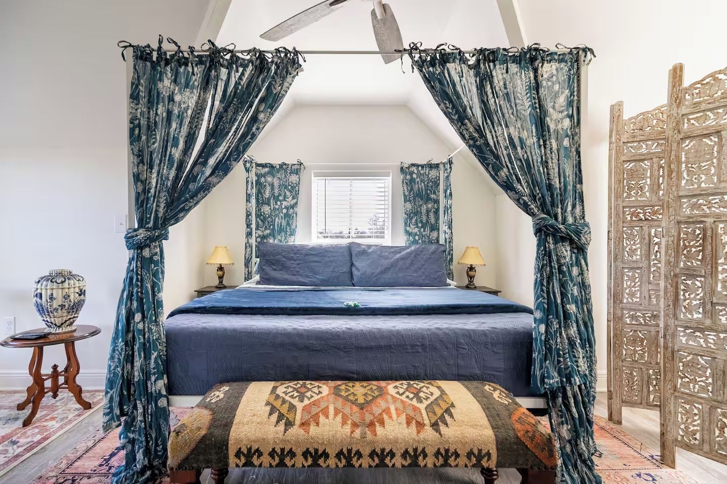 A bedroom with a canopy bed, two bedside lamps, a patterned throw pillow, and a decorative vase on a side table.