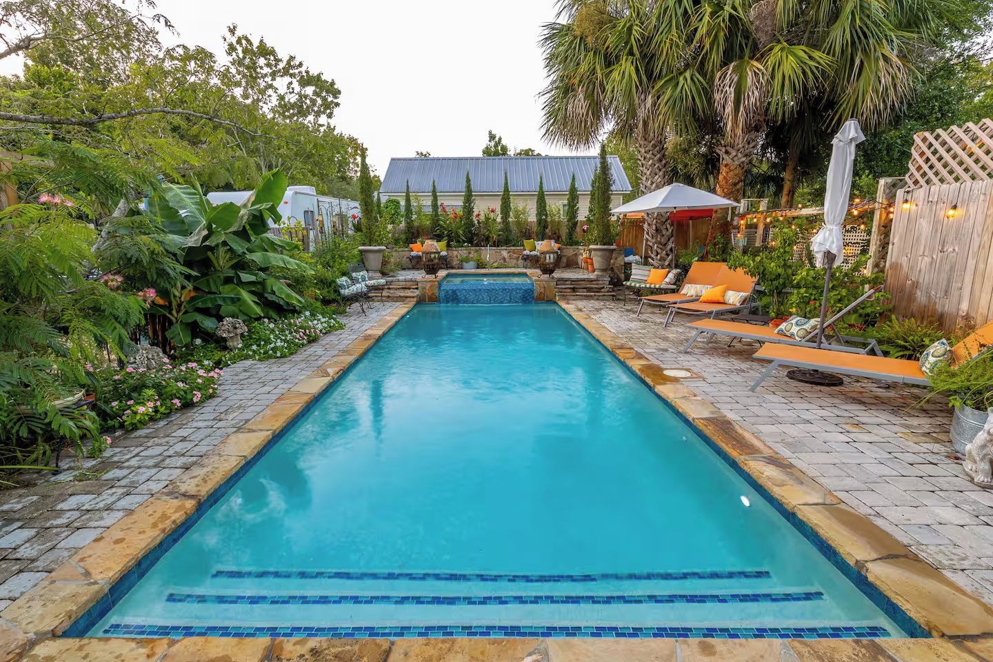 A rectangular swimming pool with a tiled edge, surrounded by a garden with various plants and a wooden fence.