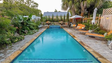 A rectangular swimming pool with a tiled edge, surrounded by a garden with various plants and a wooden fence.