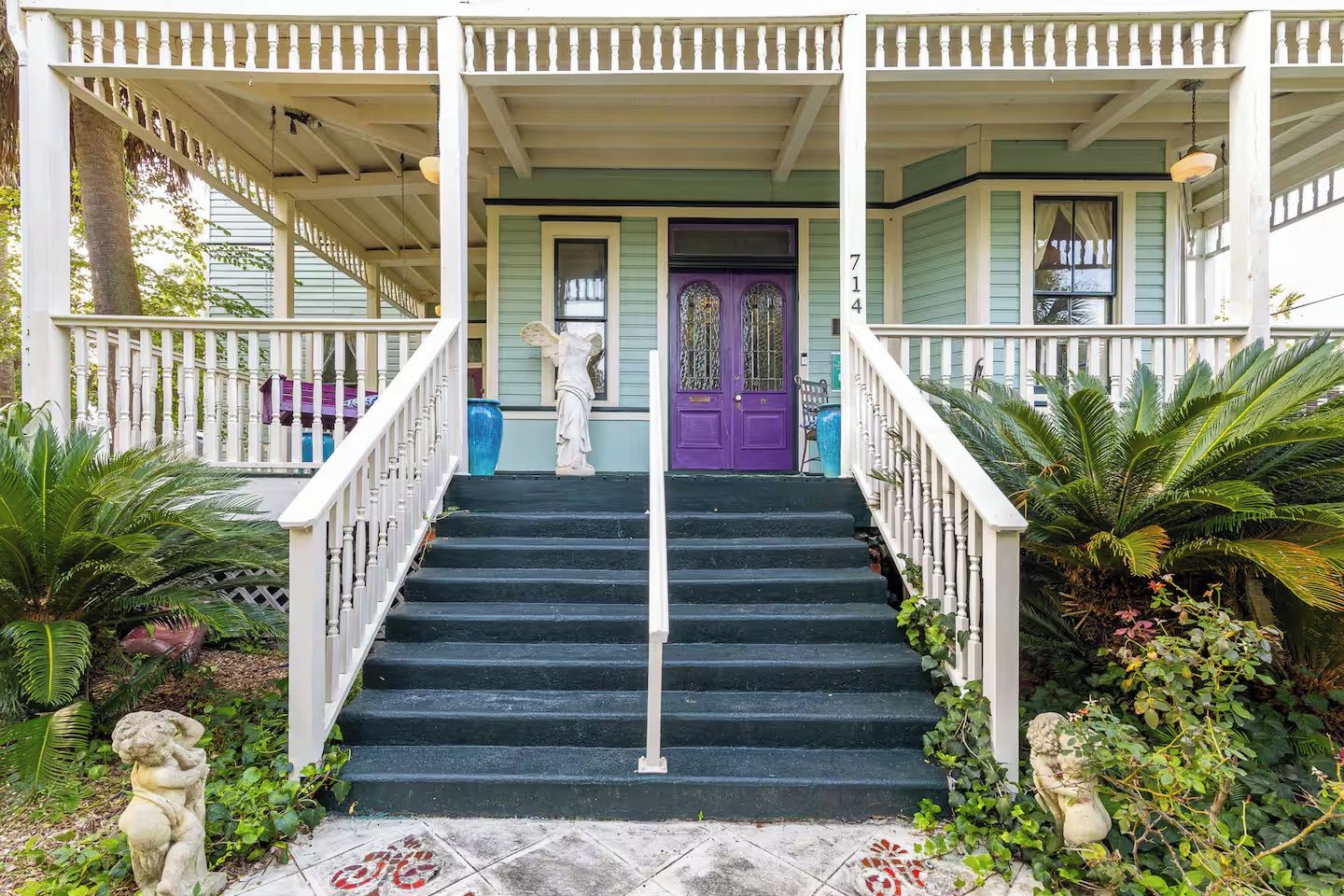 A porch with a staircase leading to a door, surrounded by plants and statues.