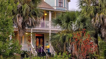 A two-story house with a balcony, surrounded by various plants and trees.