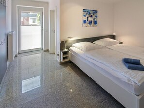 A hotel room with a bed, a nightstand, a lamp, a painting on the wall, and a door leading to the outside. - Holiday Home With 2 Bedrooms (Esens)