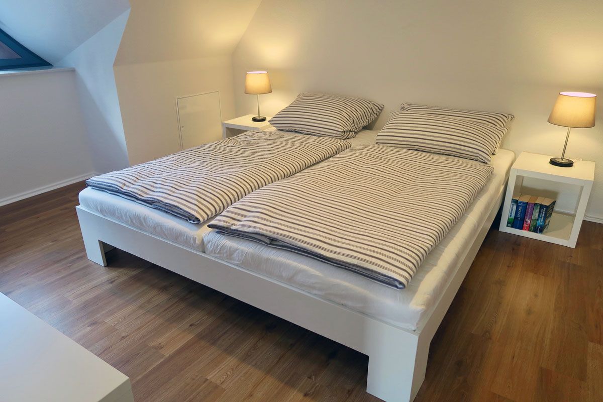 A double bed with striped bedding, two bedside tables with lamps, and a small bookshelf.