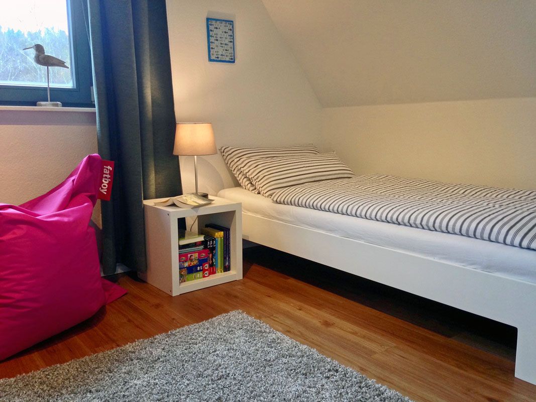 A bedroom with a bed, a nightstand with a lamp, a bookshelf, and a window with a bird decoration.