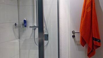 Ein Duschbereich mit Glastür, einem Handtuchhalter mit orangefarbenem Handtuch und einer kleinen Ablage mit Pflegeprodukten.