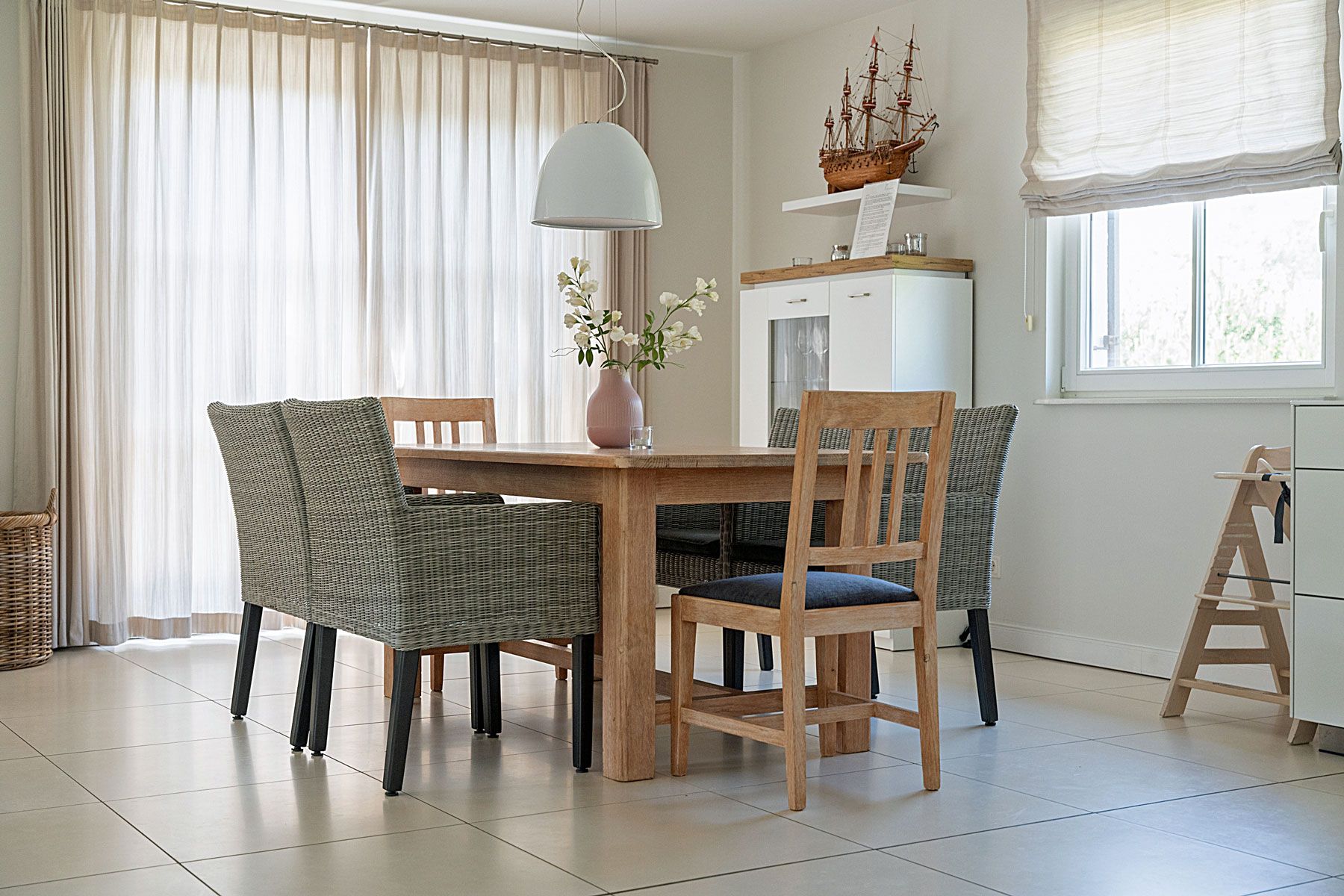 Uma sala de jantar com mesa de madeira, cadeiras de vime, um vaso com flores e um modelo de navio em uma estante.