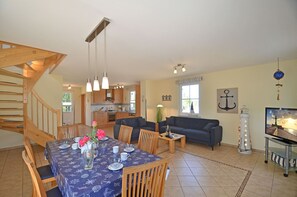 Amplia sala de estar con una mesa de comedor preparada para cuatro personas, un sofá y un televisor. Una escalera de madera conduce a un nivel superior.