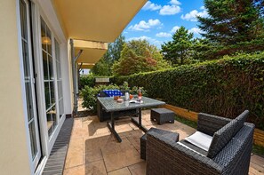 Een terras buiten met een tafel en rieten meubels, een haag en een heldere lucht.