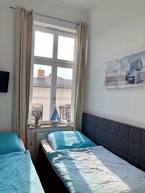 Quarto com duas camas, janela com cortinas, radiador, TV fixada na parede e pintura com tema náutico.