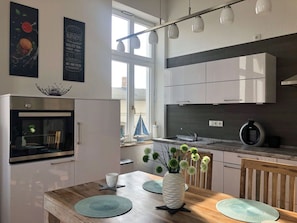 Eine moderne Küche mit weißen Schränken, einem eingebauten Backofen und einem Essbereich mit einem Holztisch und Unterlagen.