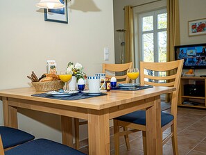 Ein Esstisch ist gedeckt mit Frühstücksangeboten, zwei Stühle stehen daneben, im Hintergrund ein Fernseher und ein Fenster mit Vorhängen.
