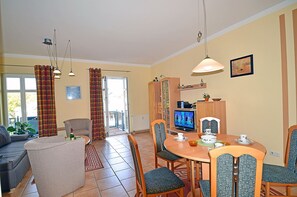 A living room with a sofa, a dining table with chairs, a television, and a kitchen area with cabinets. - Just 100 meters from the sandy Baltic Sea beach, Wi-Fi, 1 bedroom, sleeps up to 4, with a balcony (Glowe)