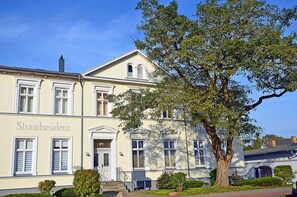 Un immeuble de deux étages avec une enseigne « Strandresidenz » sur la façade, un grand arbre devant et un ciel bleu dégagé.