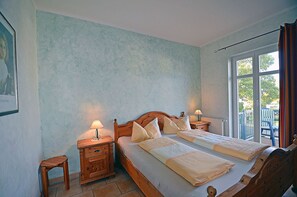 Un lit double avec une tête de lit en bois, deux tables de chevet avec des lampes, un petit tabouret en bois et une vue sur l’extérieur par une fenêtre.