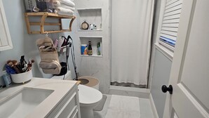 Baño con inodoro, lavamanos y zona de ducha. Hay repisas con artículos de aseo personal y un perchero.