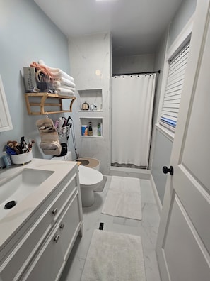 Casa de banho com sanita, lavatório e zona de duche. Existem prateleiras com artigos de higiene pessoal e um porta-toalhas.