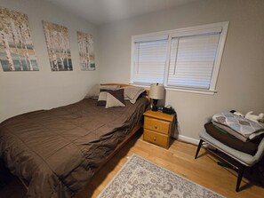 Quarto com cama, mesa de cabeceira, abajur, cadeira e janela com persianas.