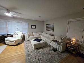 Sala de estar com sofá em formato de L, uma poltrona branca, uma mesinha, abajur e tapete.