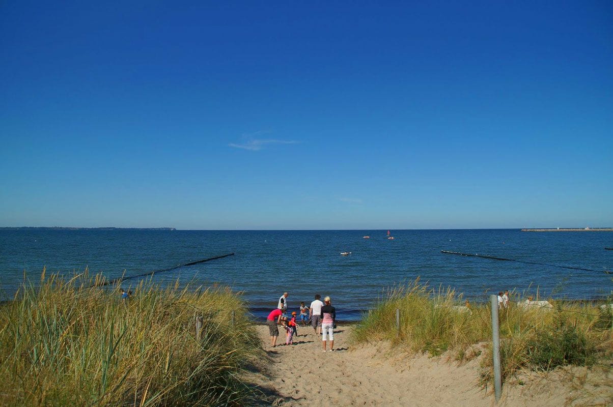 Ein sandiger Strand mit blauem Himmel, ruhiger See und einigen Personen, die am Ufer entlanggehen.
