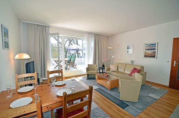 A living room with a dining table, chairs, a sofa, and a coffee table. There is a television, a lamp, and a window with curtains. - Just 250 meters from the sandy Baltic Sea beach, balcony with patio furniture and a beach umbrella (Glowe)