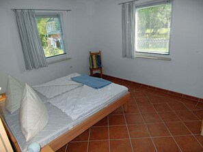 Een eenpersoonsbed met houten frame, wit beddengoed en een blauwe deken. Aan weerszijden van het bed bevinden zich twee ramen met gordijnen.