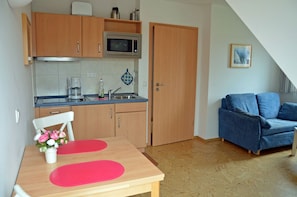 A compact kitchen with a microwave, coffee maker, and a sink. There's a dining table with red placemats and a vase of flowers. A blue sofa is in the corner, and a door leads to another room. - Only 500 meters from the sandy Baltic Sea beach, balcony with patio furniture, internet available (Glowe)