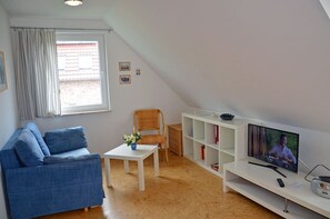 A living room with a blue sofa, a white table with a vase, a television on a white stand, and a window with curtains. - Only 500 meters from the sandy Baltic Sea beach, balcony with patio furniture, internet available (Glowe)