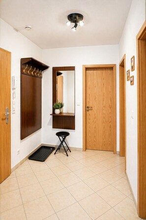 Um corredor com porta de madeira, um porta-roupas, um banquinho pequeno, um espelho e um lustre.
