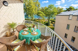 A balcony with a table set for tea, wicker chairs, and a view of a building and trees. - 2nd floor with ocean view, high-end amenities, just 30 m from the sandy beach (Baltic Sea) (Juliusruh)
