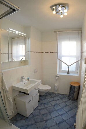 Ein Badezimmer mit einem weißen Waschbecken, einer Toilette und einem Fenster mit weißem Vorhang.