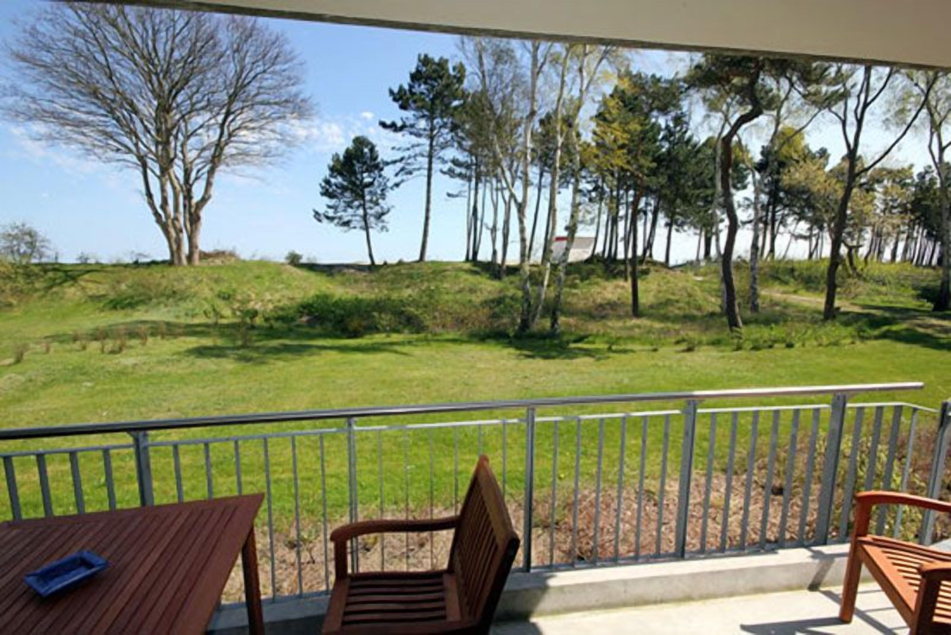 A balcony with a view of a grassy area and trees, a table, and two chairs.