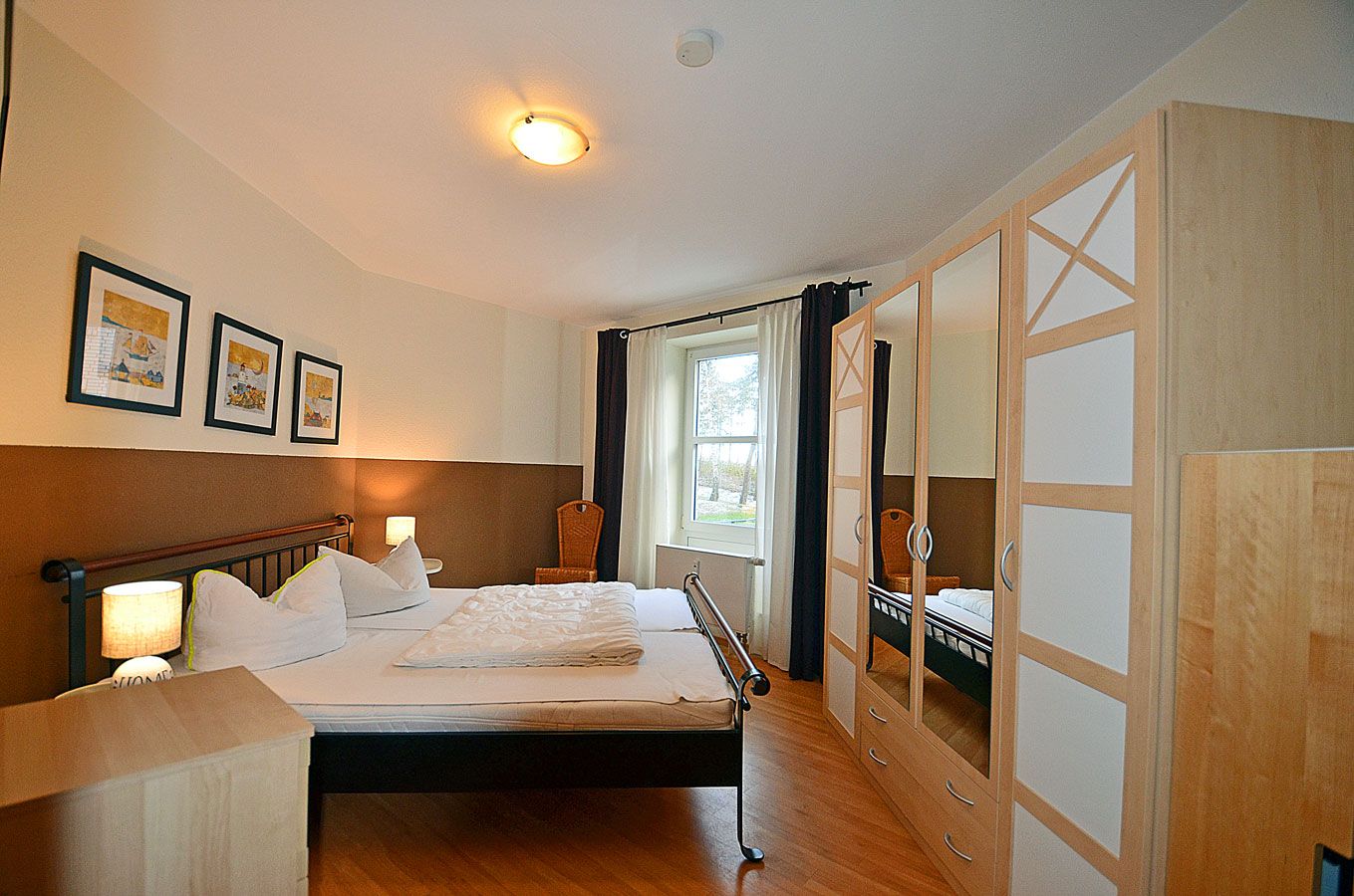 A hotel room with a bed, bedside lamps, a window with curtains, a wardrobe with mirrored doors, and framed pictures on the wall.