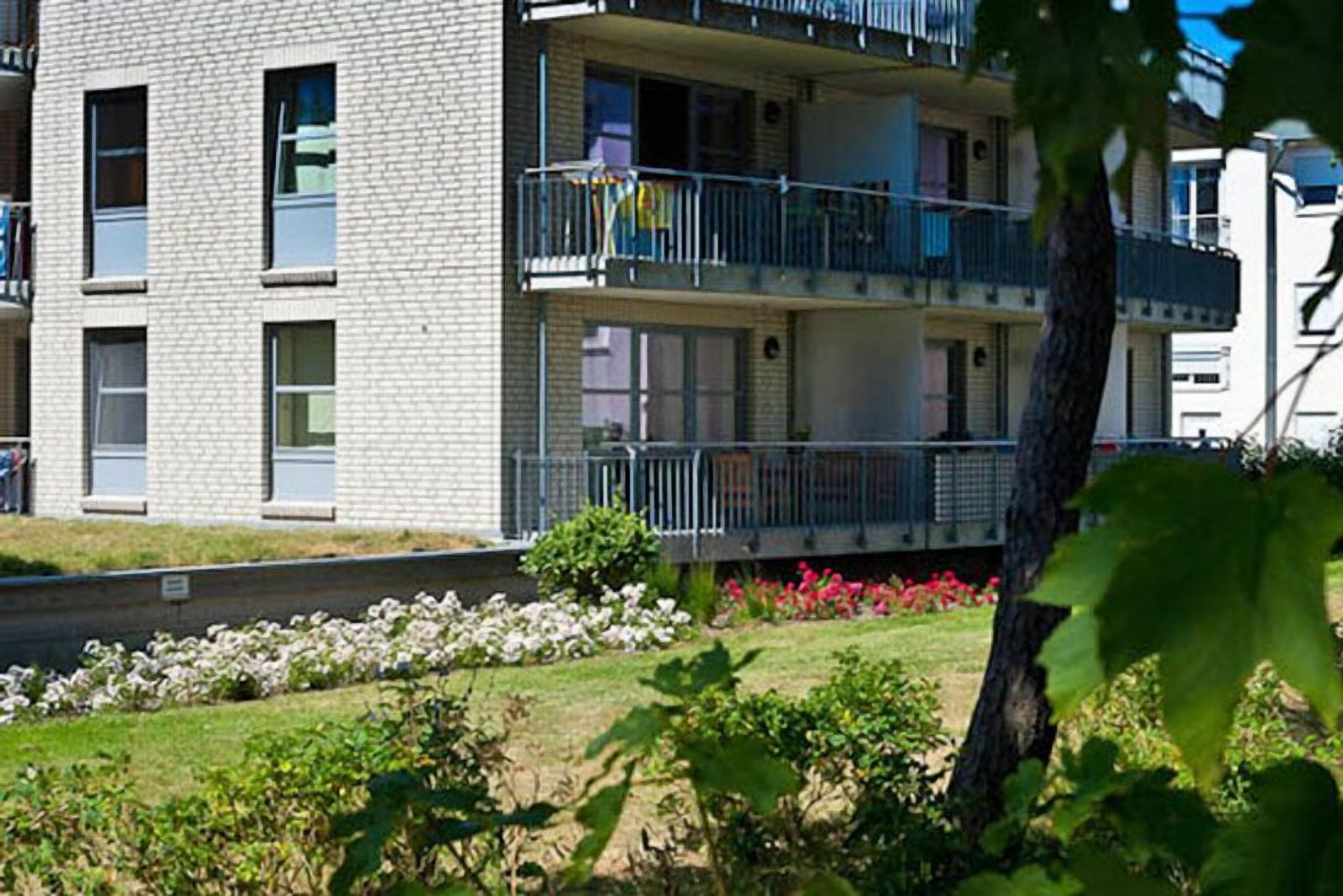A multi-story residential building with balconies, a landscaped garden, and a clear sky.