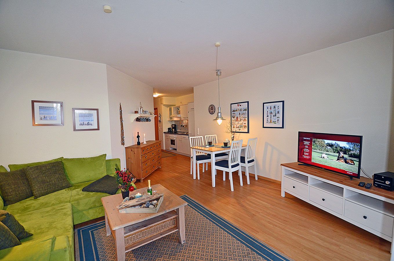 Ein Wohnzimmer mit grünem Sofa, einem Holztisch, einem Essbereich mit weißem Tisch und Stühlen, einer Küche im Hintergrund und einem Fernseher auf einem Schrank.